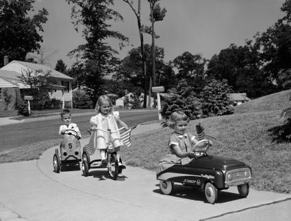 1950 niños montando coches de juguete y bicicletas en la acera